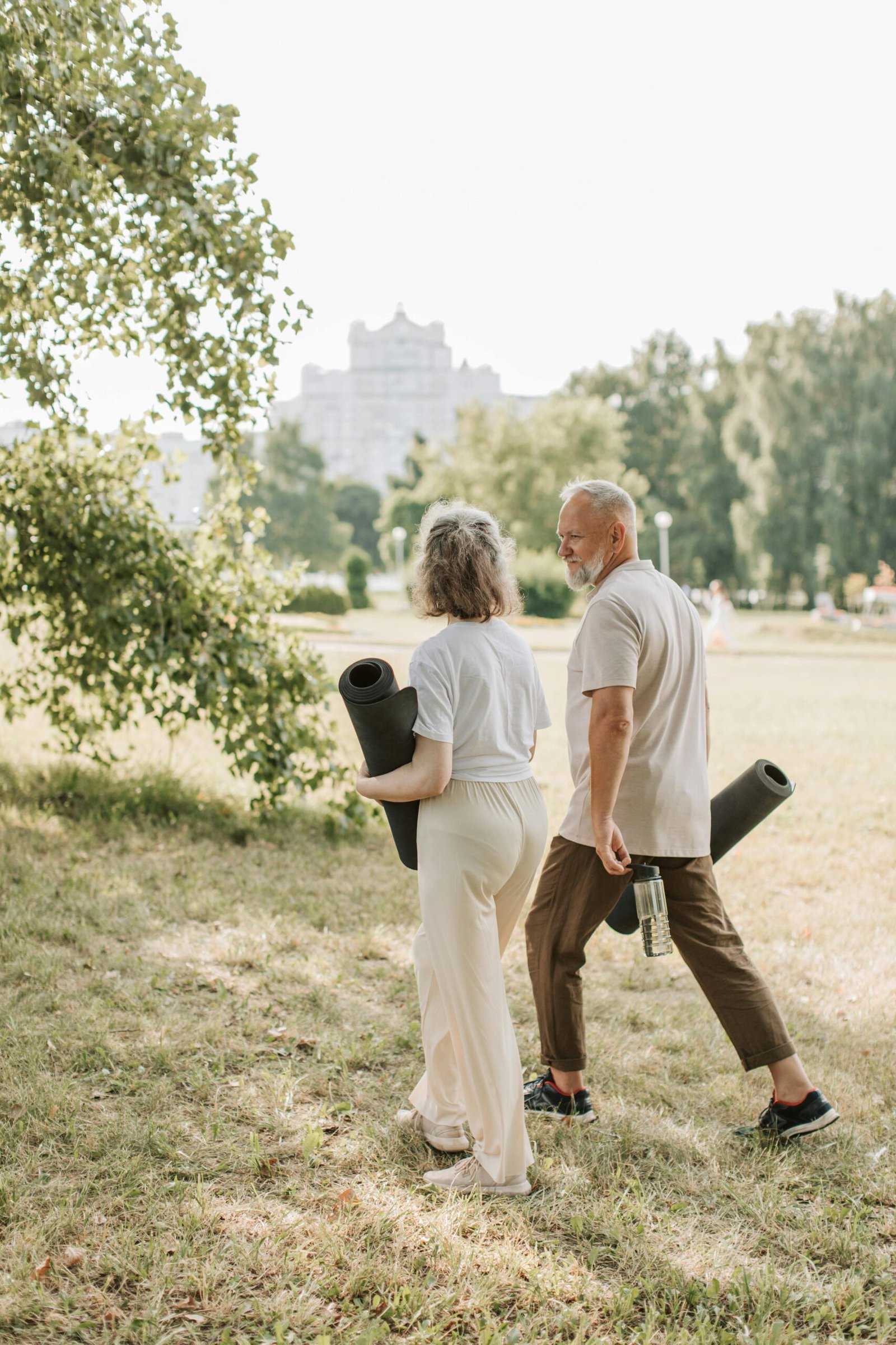 Elderly couple walking in park carrying yoga mats for a wellness session.
